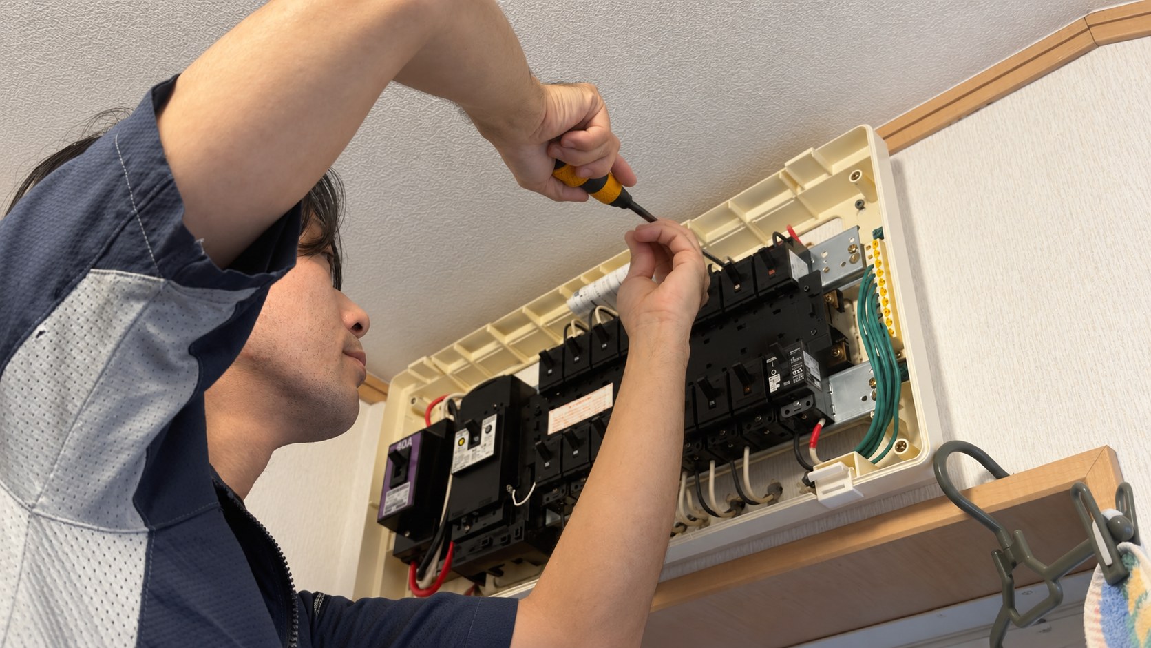 分電盤内部をテスターで確認する手元と、分電盤の配線作業を行う田島さん